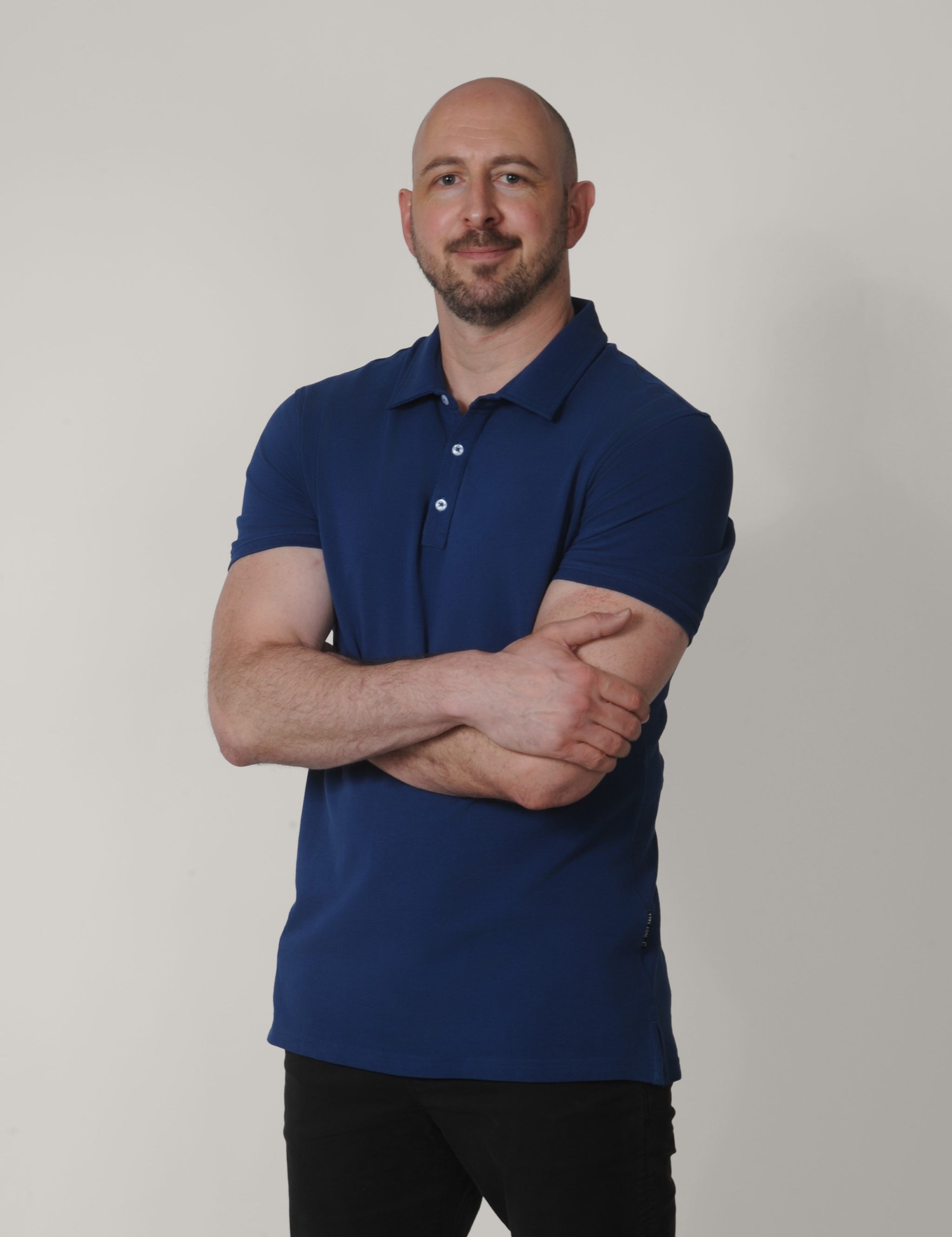 A tall and slim man in the studio standing in front of a light background with his arms crossed. The smiling model is wearing an extra long slim navy blue polo shirt in a size XL. The tall navy blue polo shirt features a 3" longer body, 100% organic cotton, and is soft & preshrunk. The navy blue polo shirt is ideal for tall slim men 6'2"+.