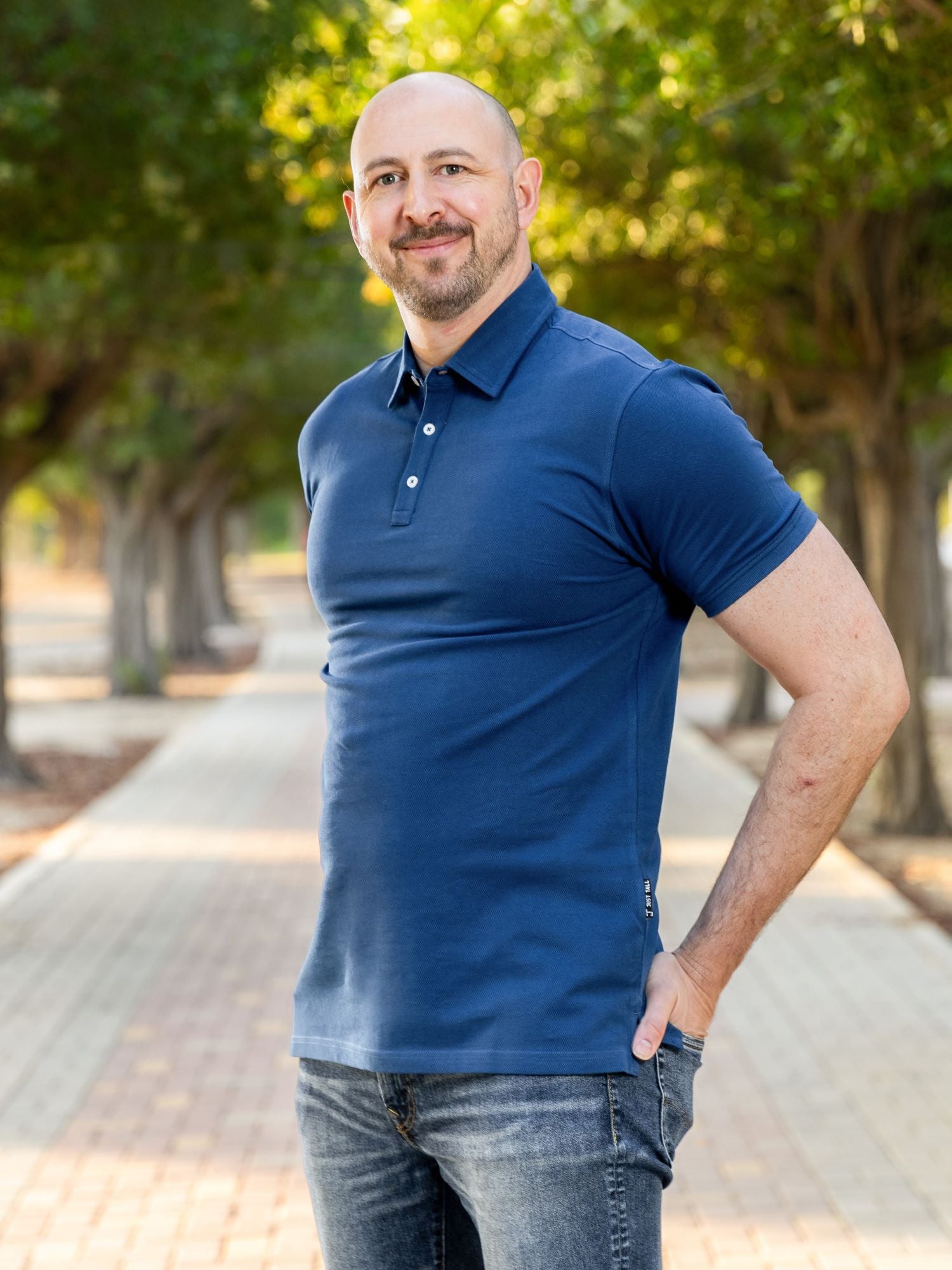 A tall athletic guy in a park with his hands in his rear pockets and wearing a navy XL tall polo shirt.