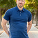 A tall and athletic man in a park standing on a path between trees. The smiling model is wearing an extra long slim navy polo shirt in a size XL. The tall navy polo shirt features a 3" longer body, 100% organic cotton, and is soft & preshrunk. The navy polo shirt is ideal for tall slim men 6'2"+.