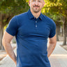 A tall and athletic man in a park standing on a path between trees. The smiling model is wearing an extra long slim navy polo shirt in a size XL. The tall navy polo shirt features a 3" longer body, 100% organic cotton, and is soft & preshrunk. The navy polo shirt is ideal for tall slim men 6'2"+.