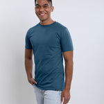 A tall and slim man in the studio standing in front of a light background with one hand in his pocket. The smiling model is wearing an extra long slim petrol t-shirt in a size medium. The tall petrol t-shirt features a 3" longer body, 100% organic cotton, and is soft & preshrunk. The petrol t-shirt is ideal for tall slim men 6'2"+.