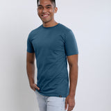 A tall and slim man in the studio standing in front of a light background with one hand in his pocket. The smiling model is wearing an extra long slim petrol t-shirt in a size medium. The tall petrol t-shirt features a 3" longer body, 100% organic cotton, and is soft & preshrunk. The petrol t-shirt is ideal for tall slim men 6'2"+.
