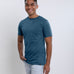 A tall and slim man in the studio standing in front of a light background with one hand in his pocket. The smiling model is wearing an extra long slim petrol t-shirt in a size medium. The tall petrol t-shirt features a 3" longer body, 100% organic cotton, and is soft & preshrunk. The petrol t-shirt is ideal for tall slim men 6'2"+.