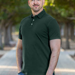 A tall and athletic man in a park standing on a path between trees. The smiling model is wearing an extra long slim dark pine polo shirt in a size XL. The tall dark pine polo shirt features a 3" longer body, 100% organic cotton, and is soft & preshrunk. The dark pine polo shirt is ideal for tall slim men 6'2"+. Buy 2 Save 10%, Buy 4 Save 15%, Buy 6 Save 20%.