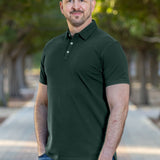 A tall and athletic man in a park standing on a path between trees. The smiling model is wearing an extra long slim dark pine polo shirt in a size XL. The tall dark pine polo shirt features a 3" longer body, 100% organic cotton, and is soft & preshrunk. The dark pine polo shirt is ideal for tall slim men 6'2"+. Buy 2 Save 10%, Buy 4 Save 15%, Buy 6 Save 20%.