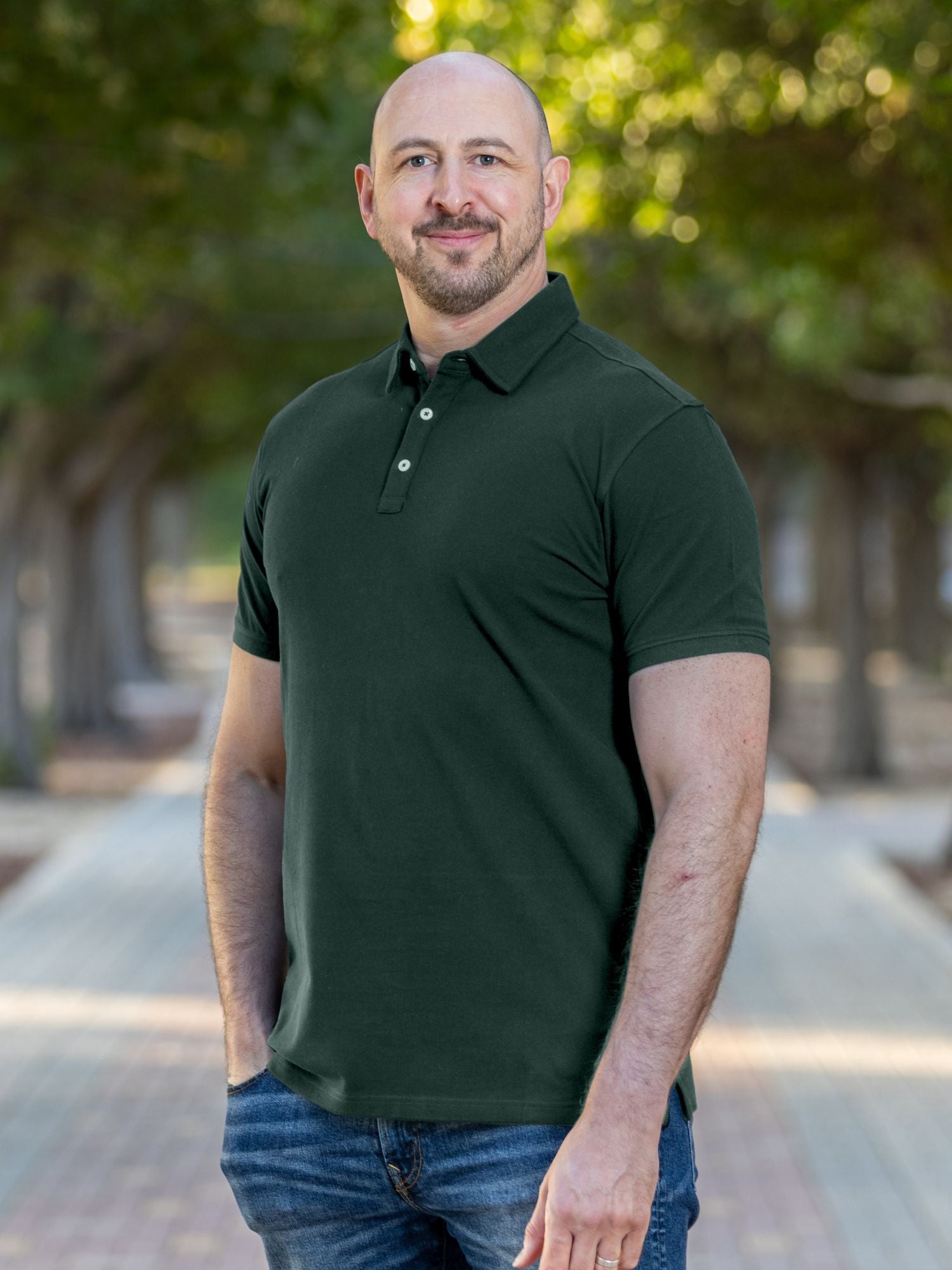 A tall and athletic man in a park standing on a path between trees. The smiling model is wearing an extra long slim dark pine polo shirt in a size XL. The tall dark pine polo shirt features a 3" longer body, 100% organic cotton, and is soft & preshrunk. The dark pine polo shirt is ideal for tall slim men 6'2"+. Buy 2 Save 10%, Buy 4 Save 15%, Buy 6 Save 20%.