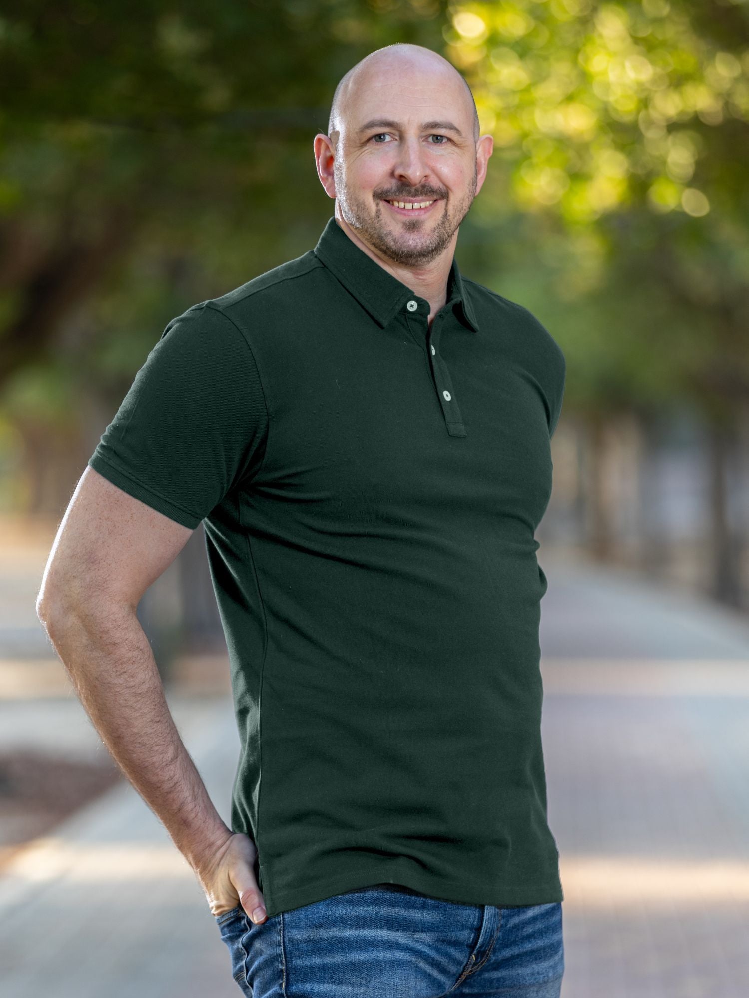 A tall and muscular man standing in a park, hands in back pockets and wearing a tall dark pine XL tall polo shirt.