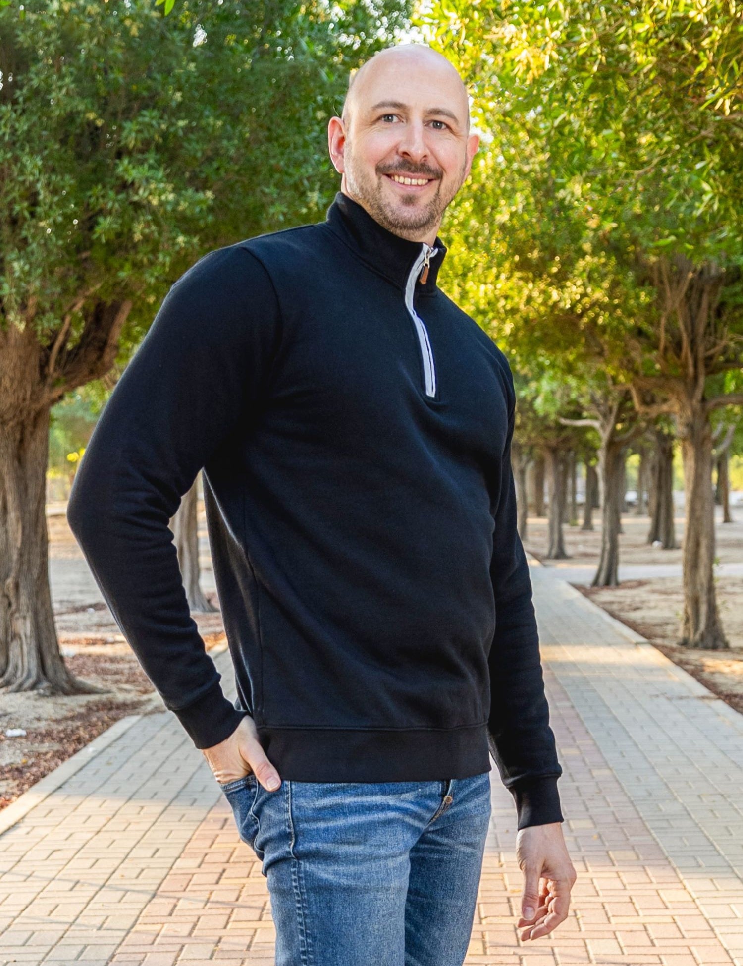 A tall and slim man standing on a path in  a park. The smiling model is wearing a black quarter zip sweatshirt. The tall black quarter zip sweatshirt features 2.5" longer sleeves, 100% organic french terry cotton, and is soft & comfy. It's  ideal for tall slim men 6'2"+.