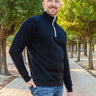 A tall and slim man standing on a path in  a park. The smiling model is wearing a black quarter zip sweatshirt. The tall black quarter zip sweatshirt features 2.5" longer sleeves, 100% organic french terry cotton, and is soft & comfy. It's  ideal for tall slim men 6'2"+.