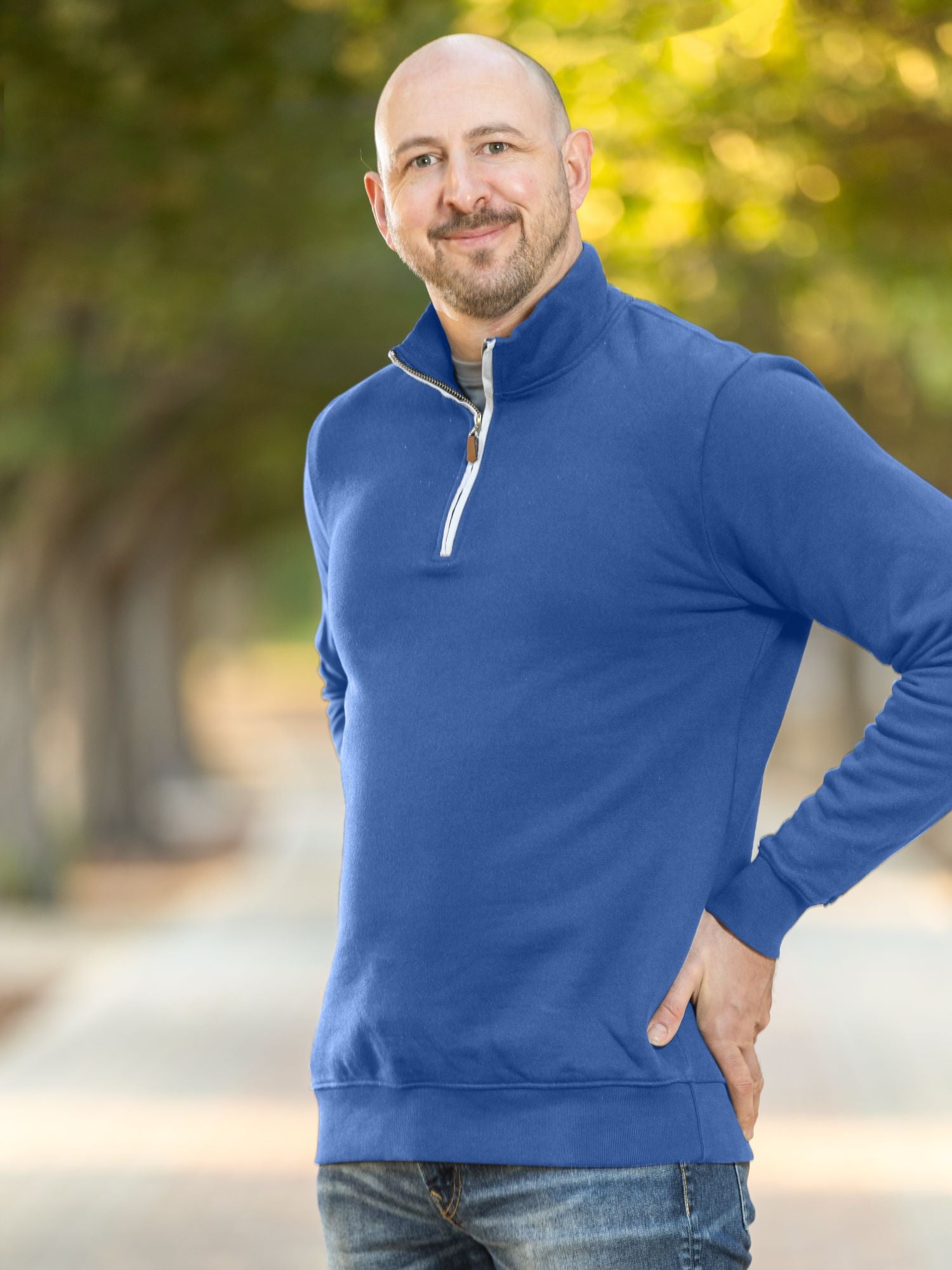 A tall and slim man standing on a path in  a park. The smiling model is wearing a cobalt quarter zip sweatshirt. The tall cobalt quarter zip sweatshirt features 2.5" longer sleeves, 100% organic french terry cotton, and is soft & comfy. It's  ideal for tall slim men 6'2"+.