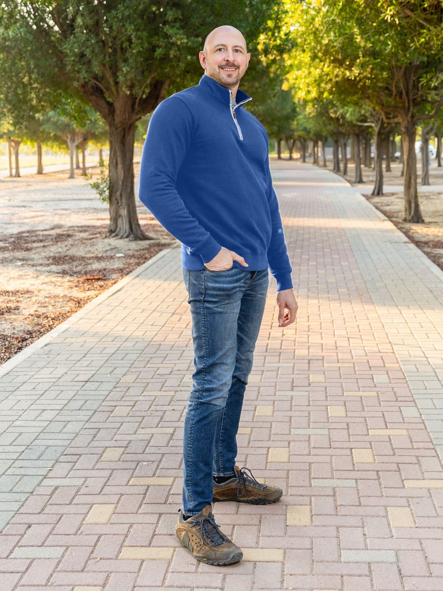 A tall slim guy wearing an XL tall cobalt quarter zip sweatshirt in a park with his hand in his pocket and smiling.