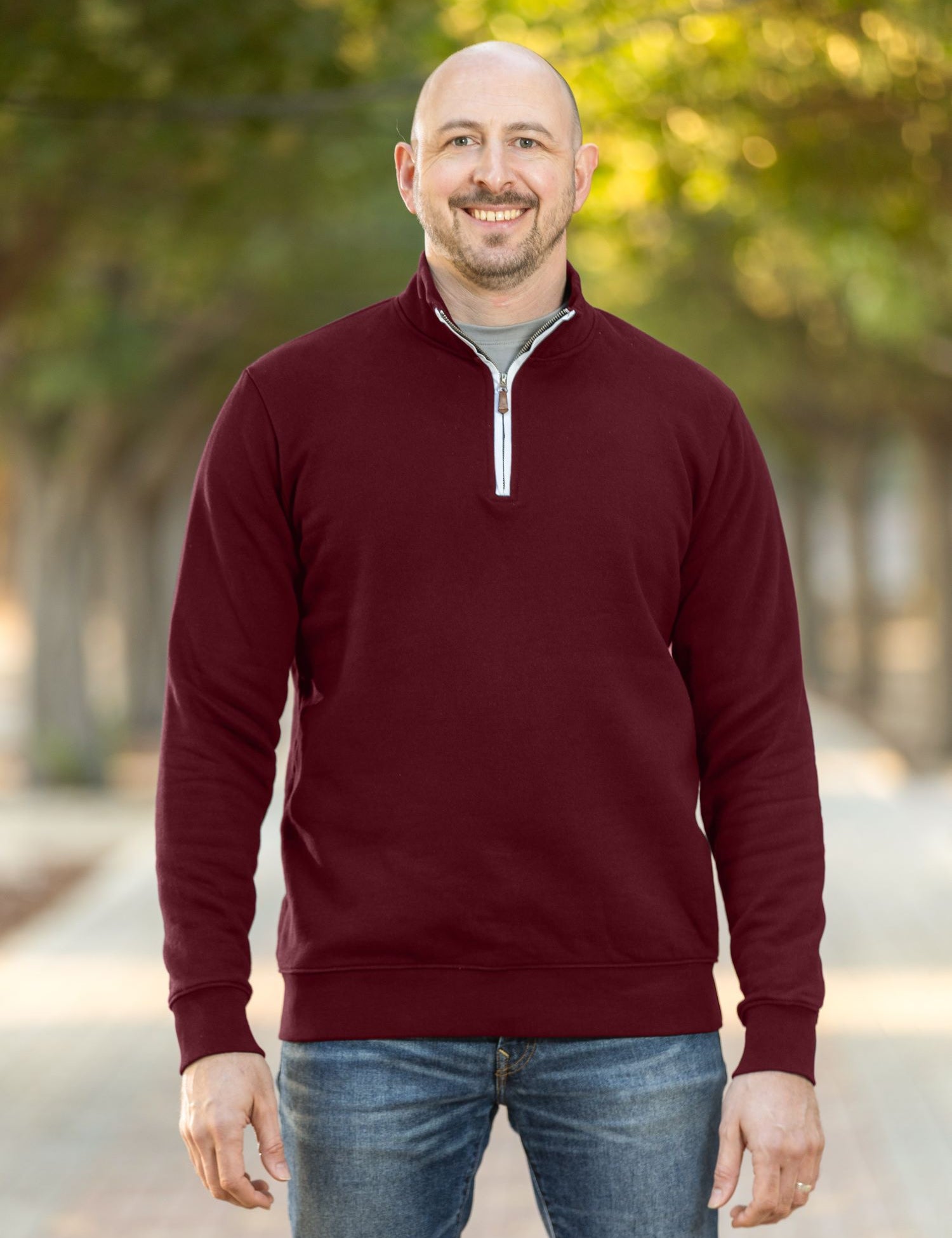 A tall and slim man standing on a path in  a park. The smiling model is wearing a maroon quarter zip sweatshirt. The tall maroon quarter zip sweatshirt features 2.5" longer sleeves, 100% organic french terry cotton, and is soft & comfy. It's  ideal for tall slim men 6'2"+.