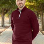 A tall slim guy wearing an XL tall maroon quarter zip sweatshirt in a park with his hands behind his smiling.