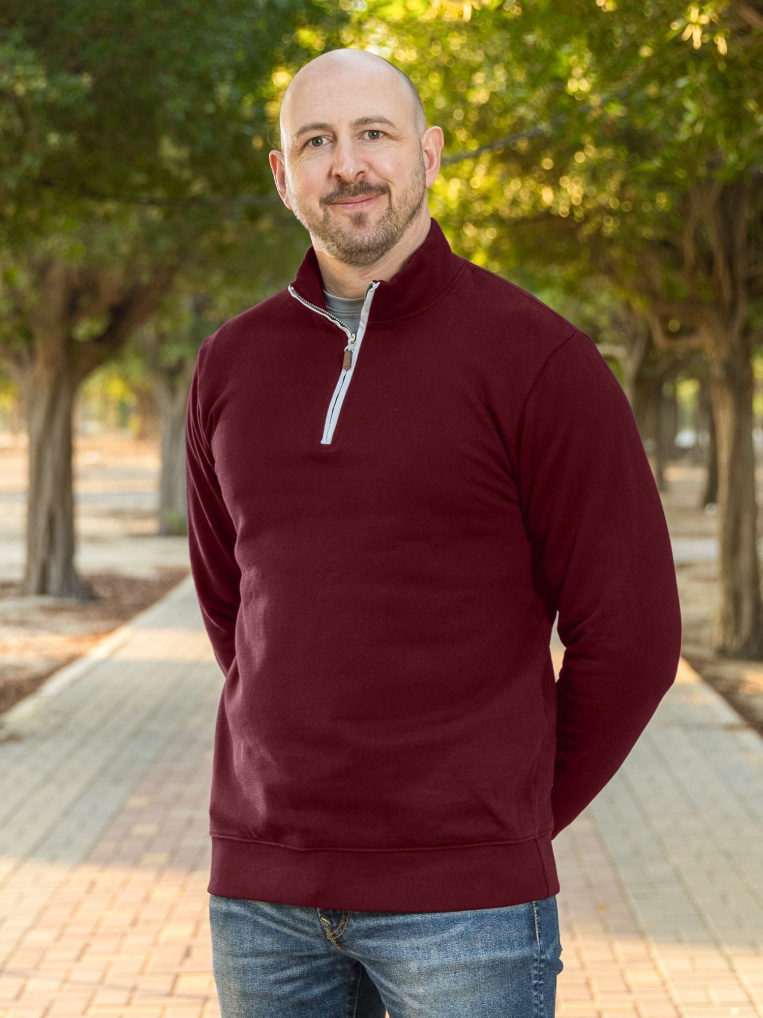 A tall slim guy wearing an XL tall maroon quarter zip sweatshirt in a park with his hands behind his smiling.