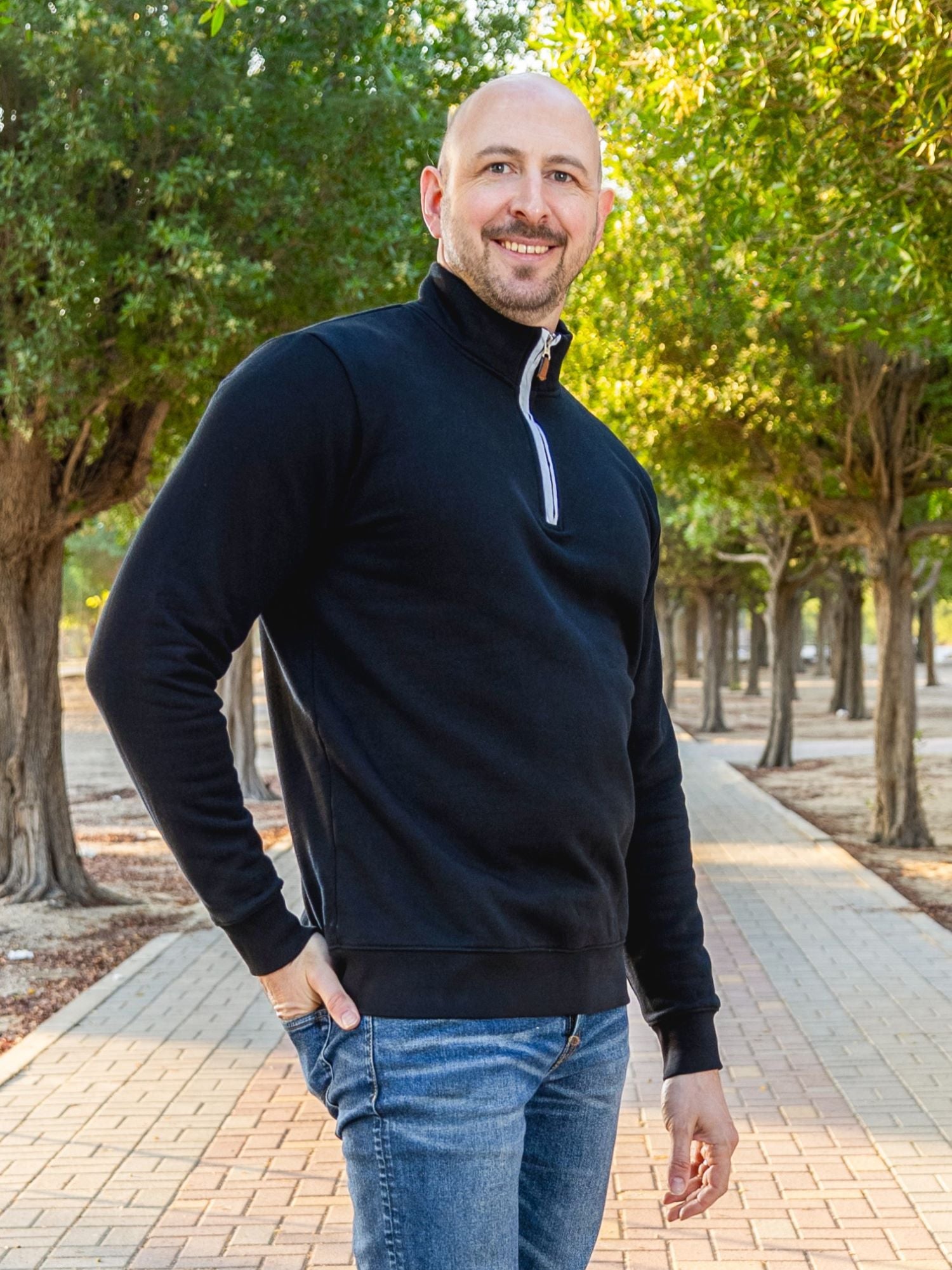 A tall and slim man standing on a path in  a park. The smiling model is wearing a black quarter zip sweatshirt. The tall black quarter zip sweatshirt features 2.5" longer sleeves, 100% organic french terry cotton, and is soft & comfy. It's  ideal for tall slim men 6'2"+.