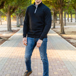 A tall slim guy wearing an XL tall black quarter zip sweatshirt in a park with his hands at his sides and smiling.