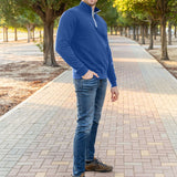 A tall slim guy wearing an XL tall cobalt quarter zip sweatshirt in a park with his hand in his pocket and smiling.