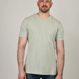 A tall and slim man in the studio standing in front of a light background. The smiling model is wearing an extra long slim sage green v-neck t-shirt in a size XL. The tall sage green v-neck t-shirt features a 3" longer body, 100% organic cotton, and is soft & preshrunk. The sage green v-neck t-shirt is ideal for tall slim men 6'2"+.