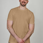 A tall athletic guy wearing a tan XL tall t-shirt, smiling and holding his left wrist with his right hand.