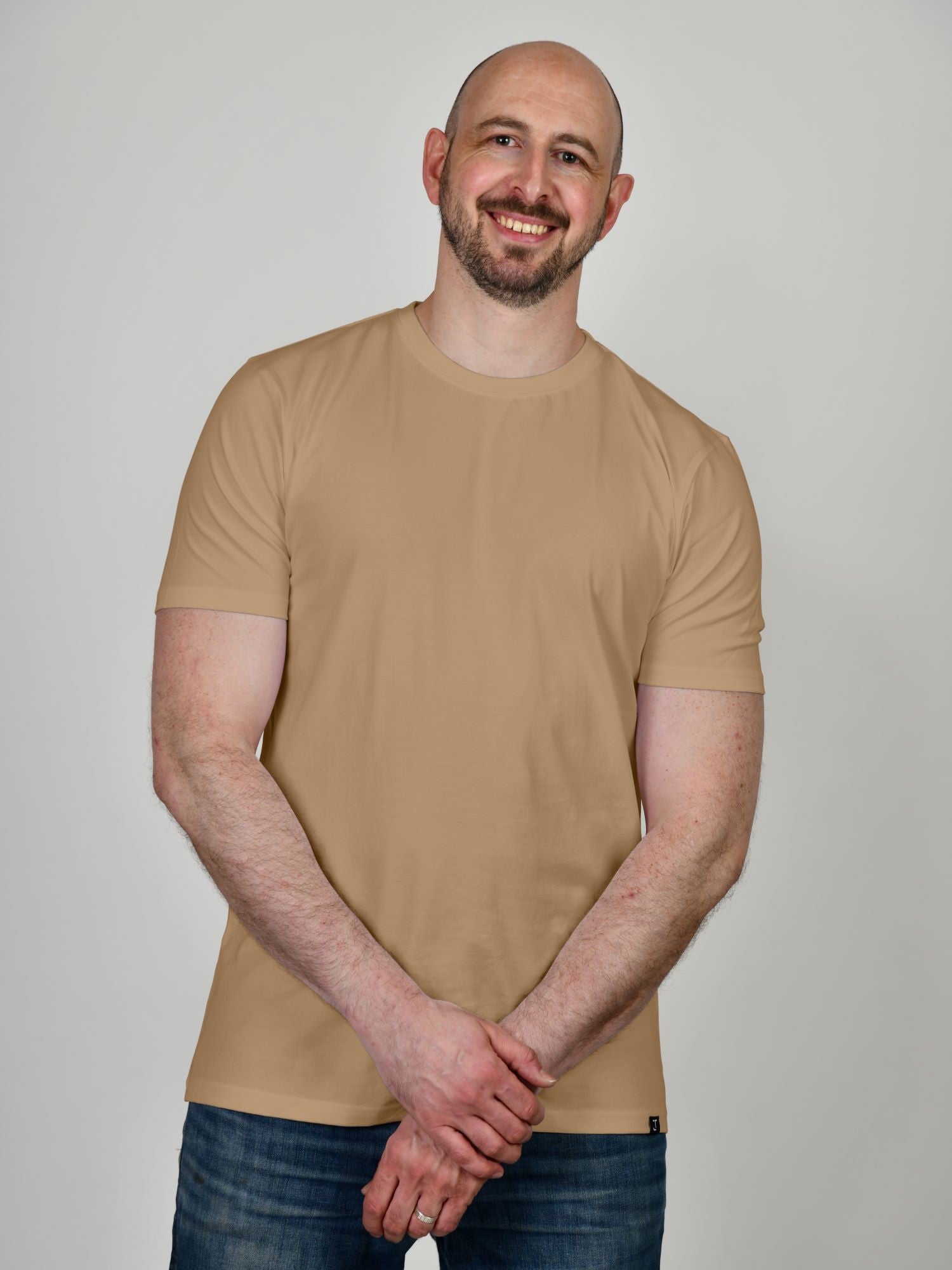 A tall athletic guy wearing a tan XL tall t-shirt, smiling and holding his left wrist with his right hand.