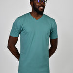 A tall and slim man in the studio standing in front of a light background with hands behind back. The smiling model is wearing an extra long slim teal v-neck t-shirt in a size large. The tall teal v-neck t-shirt features a 3" longer body, 100% organic cotton, and is soft & preshrunk. The teal v-neck t-shirt is ideal for tall slim men 6'2"+.