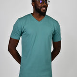 A tall and slim man in the studio standing in front of a light background with hands behind back. The smiling model is wearing an extra long slim teal v-neck t-shirt in a size large. The tall teal v-neck t-shirt features a 3" longer body, 100% organic cotton, and is soft & preshrunk. The teal v-neck t-shirt is ideal for tall slim men 6'2"+.