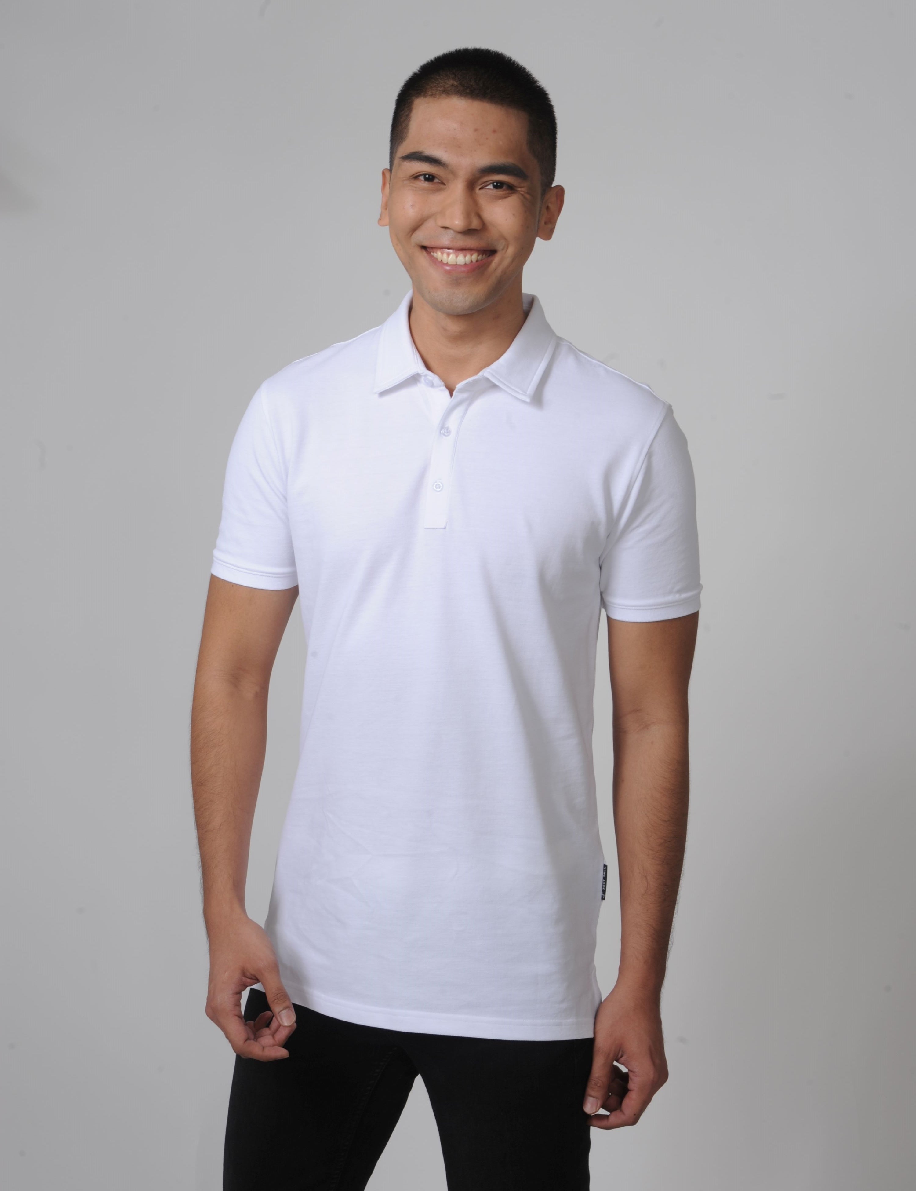 A tall and slim man in the studio standing in front of a light background. The smiling model is wearing an extra long slim white polo shirt in a size M. The tall white polo shirt features a 3" longer body, 100% organic cotton, and is soft & preshrunk. The white polo shirt is ideal for tall slim men 6'2"+.