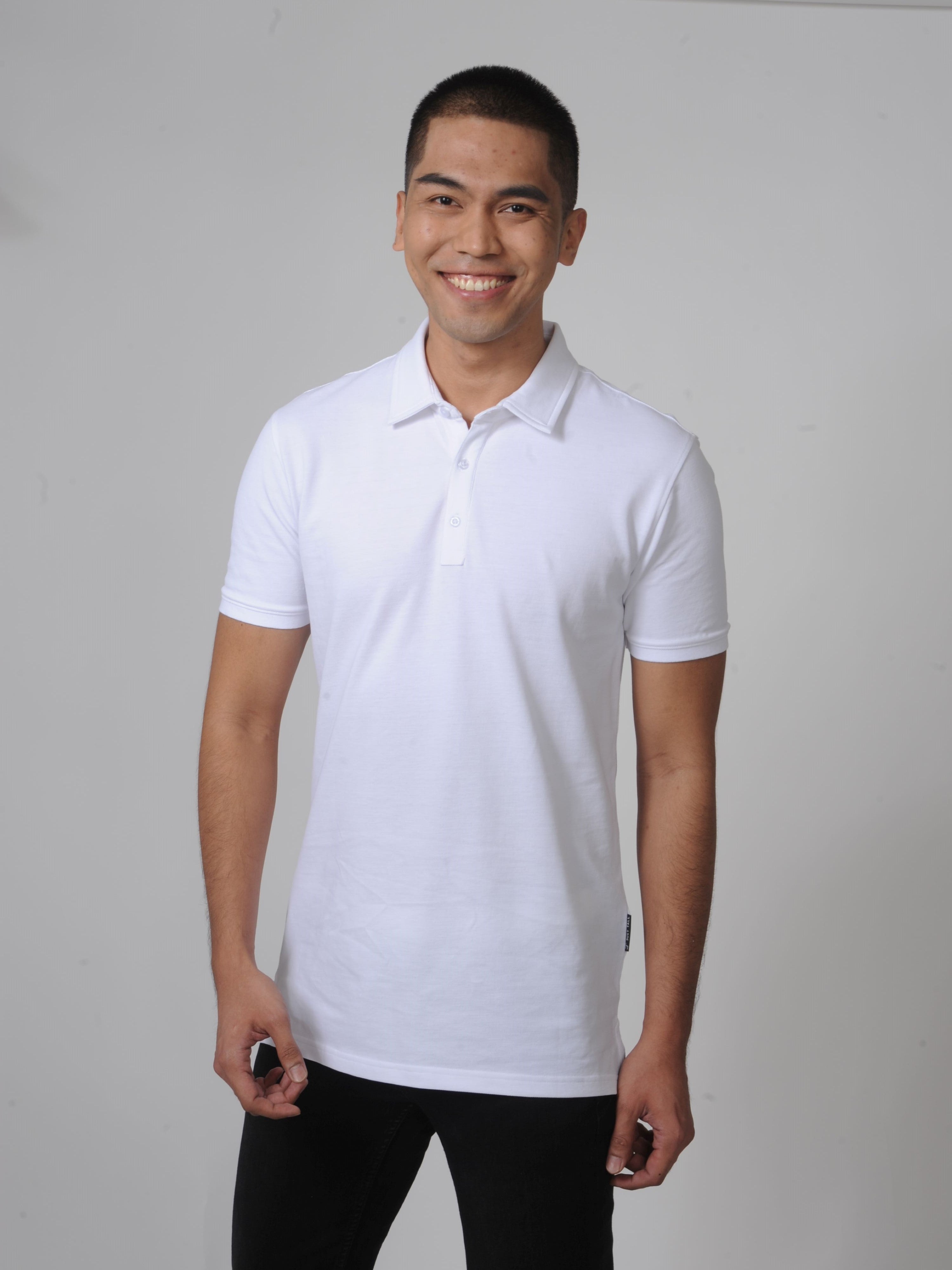 A tall and slim man in the studio standing in front of a light background. The smiling model is wearing an extra long slim white polo shirt in a size M. The tall white polo shirt features a 3" longer body, 100% organic cotton, and is soft & preshrunk. The white polo shirt is ideal for tall slim men 6'2"+.