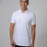 A tall and slim man in the studio standing in front of a light background. The smiling model is wearing an extra long slim white polo shirt in a size M. The tall white polo shirt features a 3" longer body, 100% organic cotton, and is soft & preshrunk. The white polo shirt is ideal for tall slim men 6'2"+.