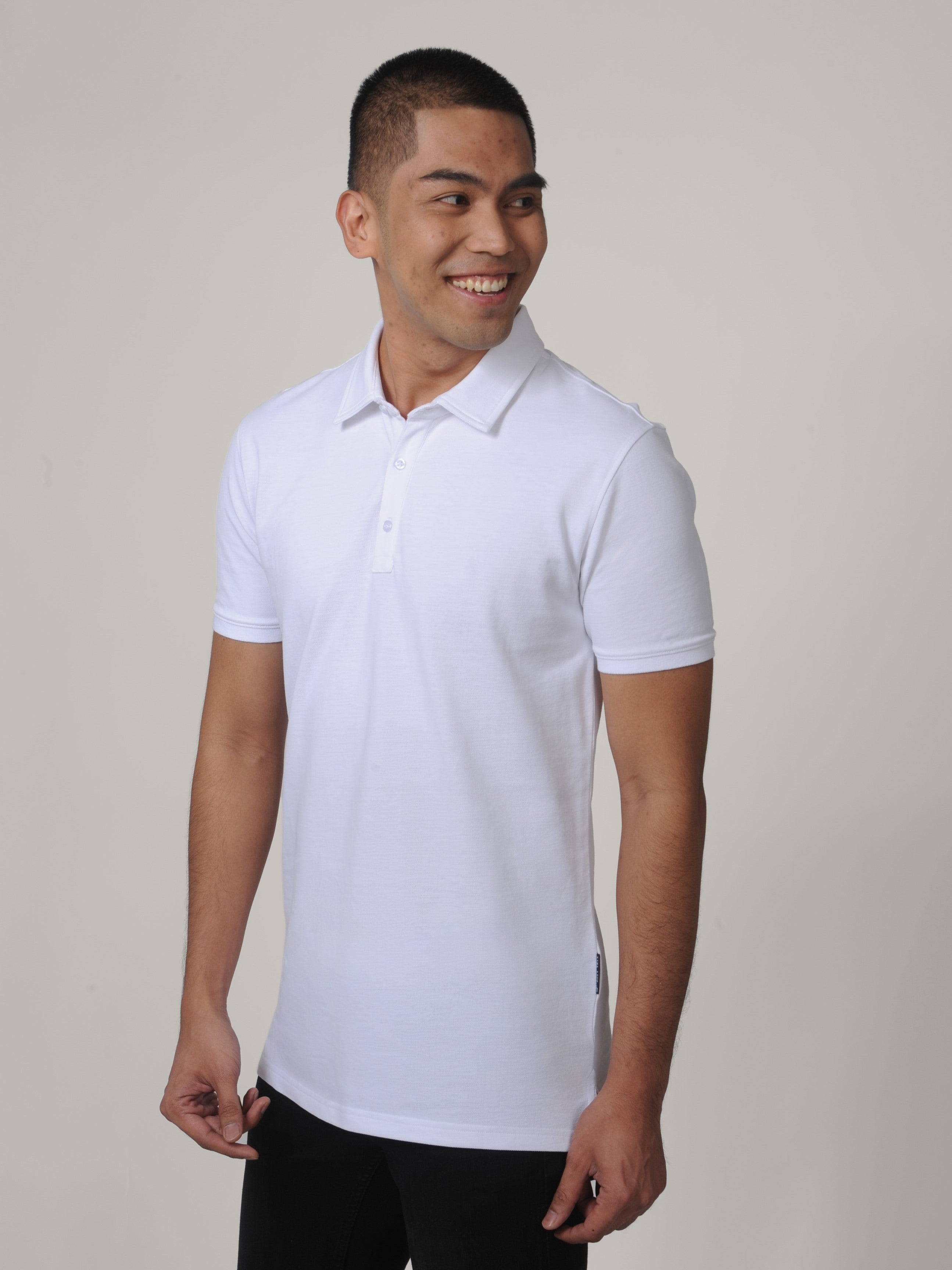A tall and slim man in the studio standing in front of a light background. The smiling model is wearing an extra long slim white polo shirt in a size medium. The tall white polo shirt features a 3" longer body, 100% organic cotton, and is soft & preshrunk. The white polo shirt is ideal for tall slim men 6'2"+.