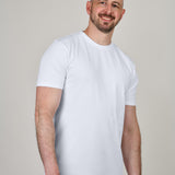 A tall and slim man in the studio standing in front of a light background. The smiling model is wearing an extra long slim white t-shirt in size a XL. The tall white t-shirt features a 3" longer body, 100% organic cotton, and is soft & preshrunk. The white t-shirt is ideal for tall slim men 6'2"+.