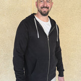 A tall slim guy standing against a wall, smiling and wearing an XL tall slim black zip up hoodie.