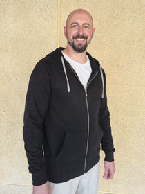 A tall slim guy standing against a wall, smiling and wearing an XL tall slim black zip up hoodie.