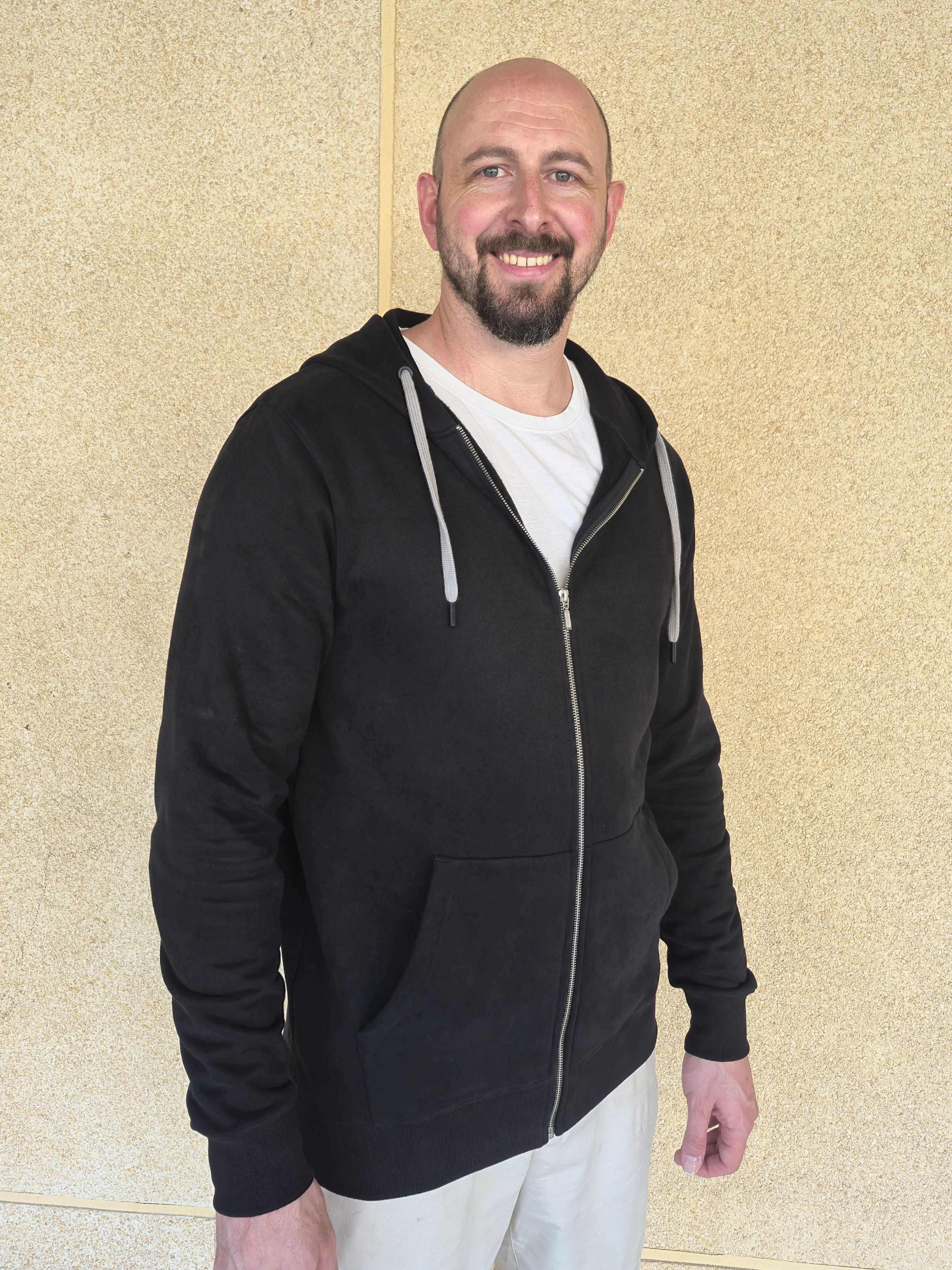 A tall slim guy standing against a wall, smiling and wearing an XL tall slim black zip up hoodie.