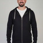 A tall slim guy standing against a wall, smiling and wearing an XL tall slim black zip up hoodie.