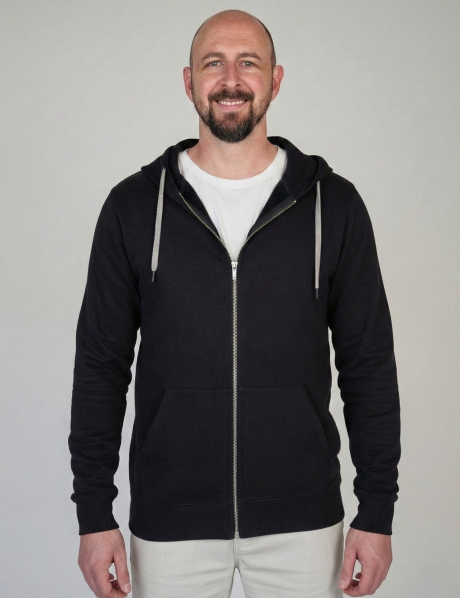 A tall slim guy standing against a wall, smiling and wearing an XL tall slim black zip up hoodie.