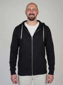 A tall slim guy standing against a wall, smiling and wearing an XL tall slim black zip up hoodie.