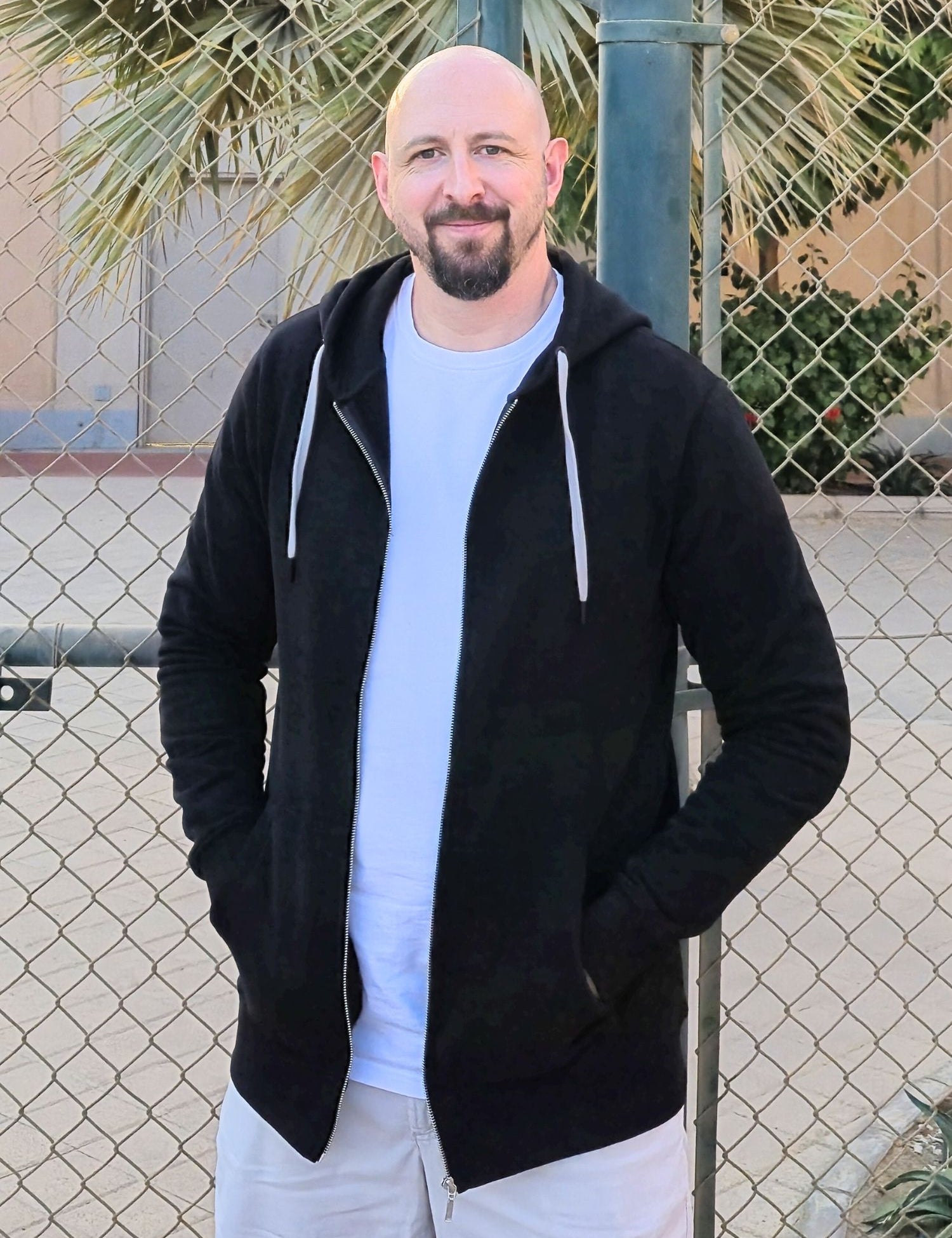 A tall slim guy standing in a tennis court, smiling and wearing an XL tall slim black zip up hoodie.