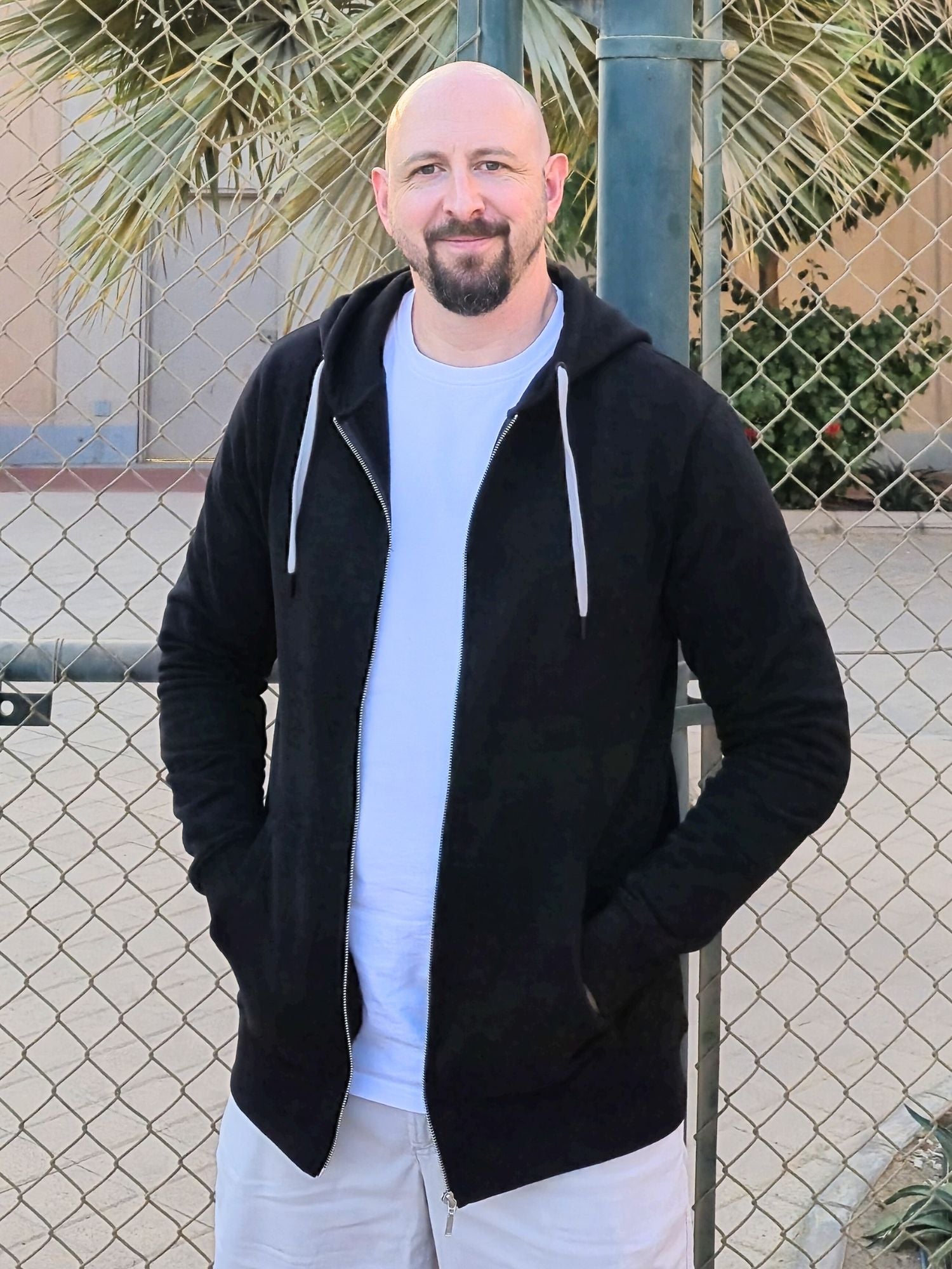 A tall slim guy standing in a tennis court, smiling and wearing an XL tall slim black zip up hoodie.