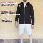 A head to toe shot of a tall slim guy standing in front of a beige wall, smiling and wearing an XL tall slim black zip up hoodie.