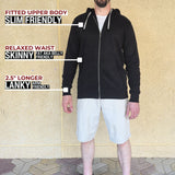 A head to toe shot of a tall slim guy standing in front of a beige wall, smiling and wearing an XL tall slim black zip up hoodie.