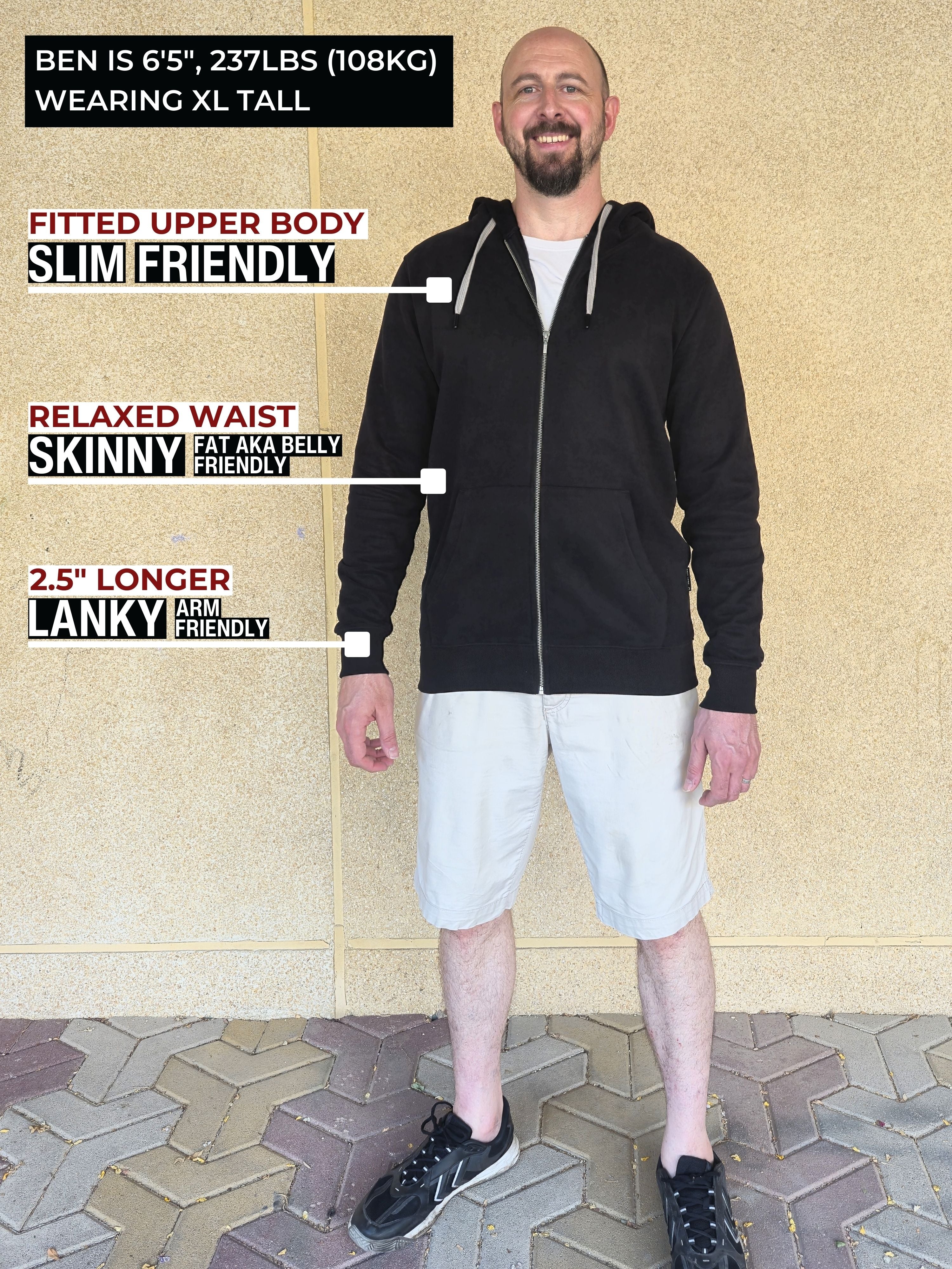 A head to toe shot of a tall slim guy standing in front of a beige wall, smiling and wearing an XL tall slim black zip up hoodie.