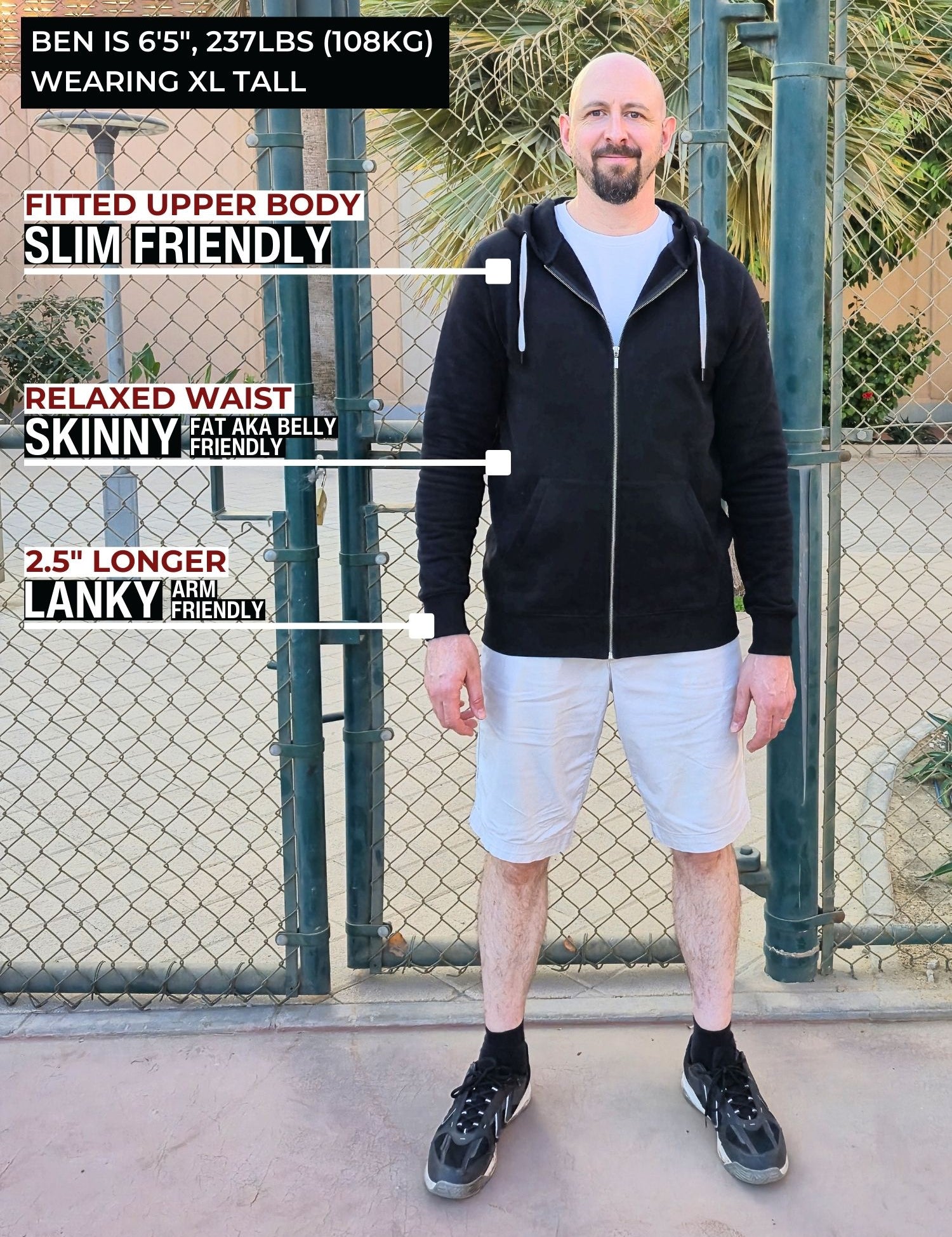 A head to toe shot of a tall slim guy standing in a tennis court, smiling and wearing an XL tall slim black zip up hoodie.