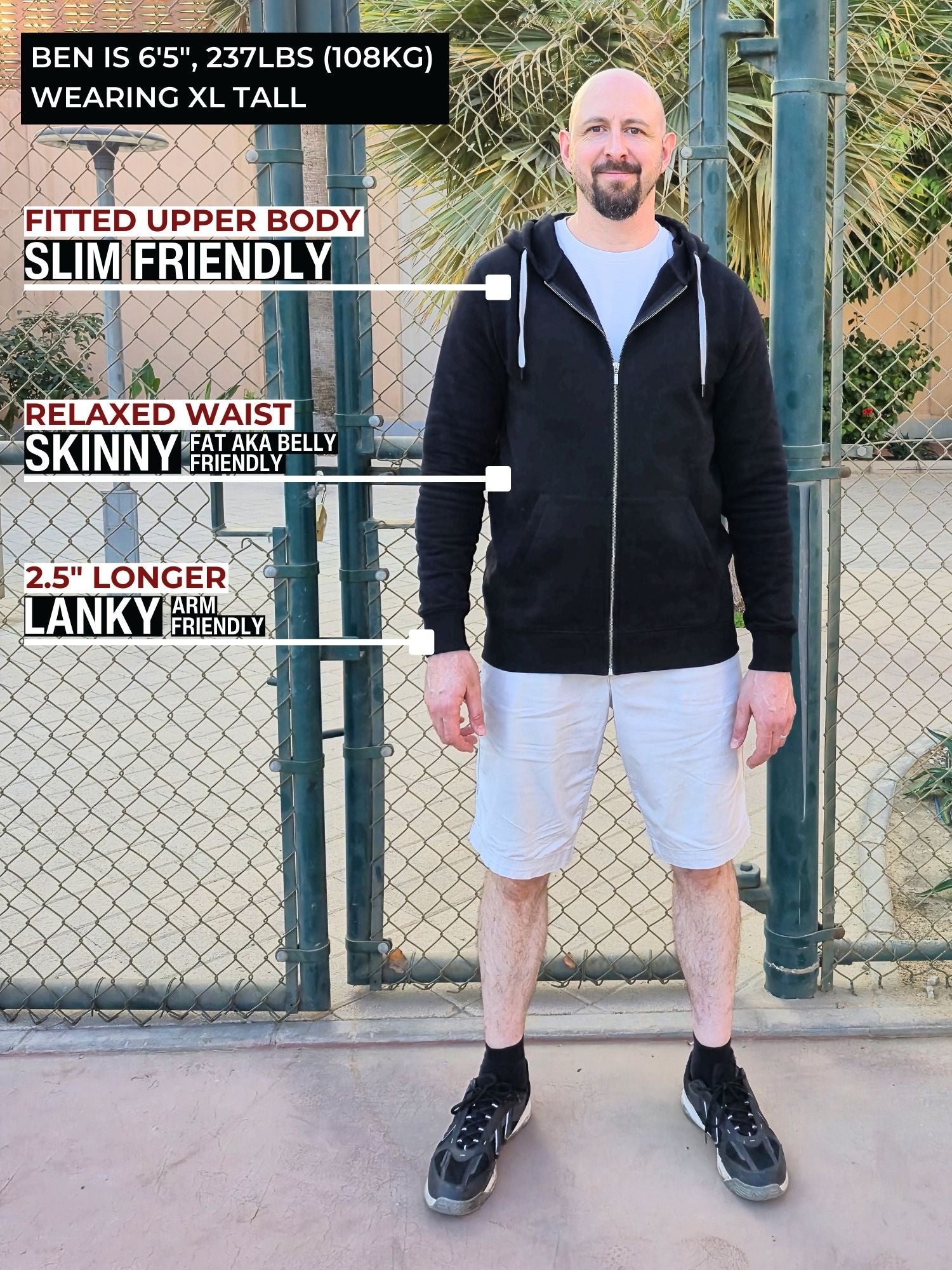 A head to toe shot of a tall slim guy standing in a tennis court, smiling and wearing an XL tall slim black zip up hoodie.