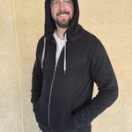 A tall slim guy wearing an XL tall black hoodie, looking to the left and hood on, standing against a beige wall.