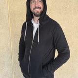 A tall slim guy wearing an XL tall black hoodie, looking to the left and hood on, standing against a beige wall.