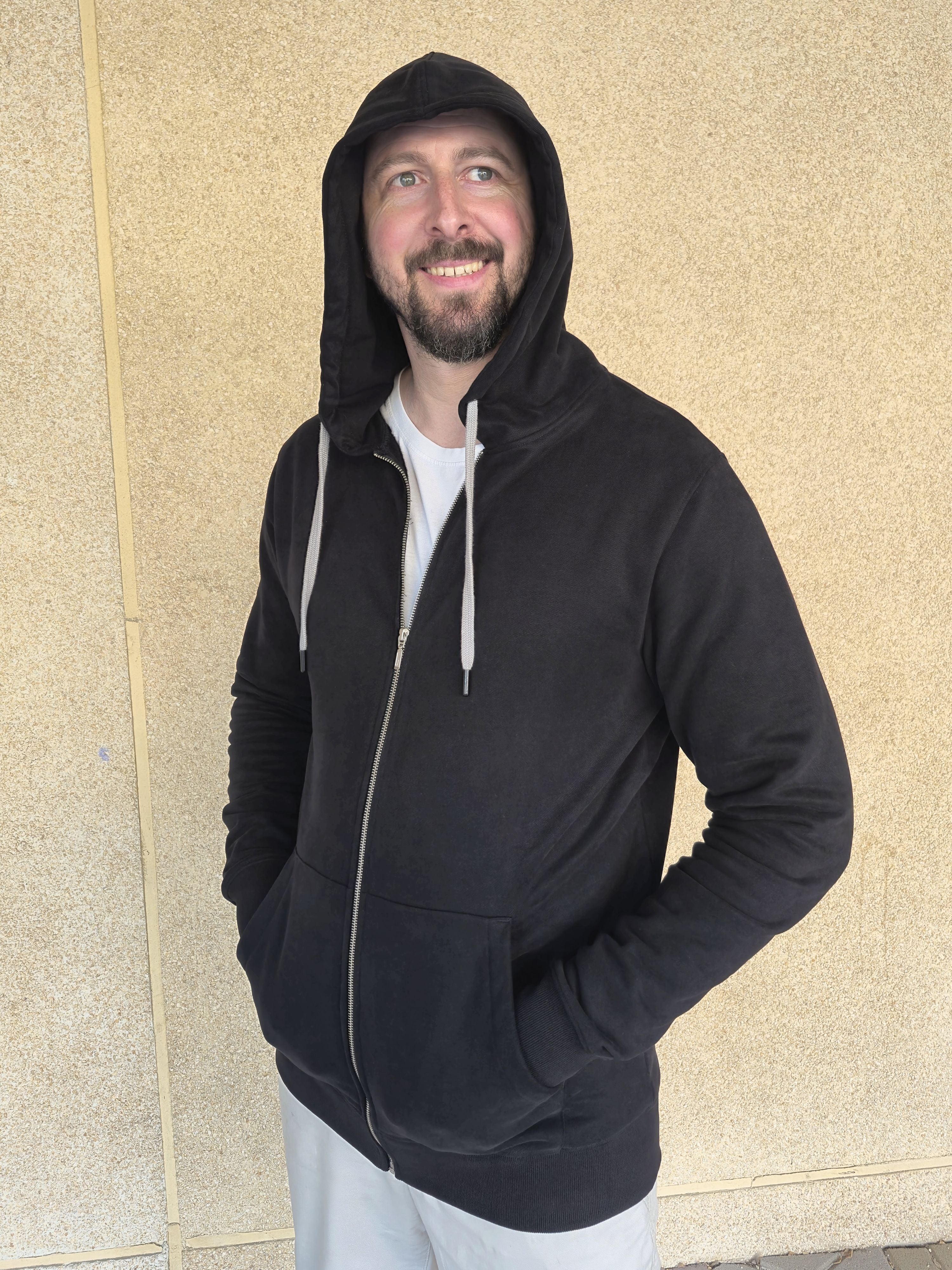 A tall slim guy wearing an XL tall black hoodie, looking to the left and hood on, standing against a beige wall.