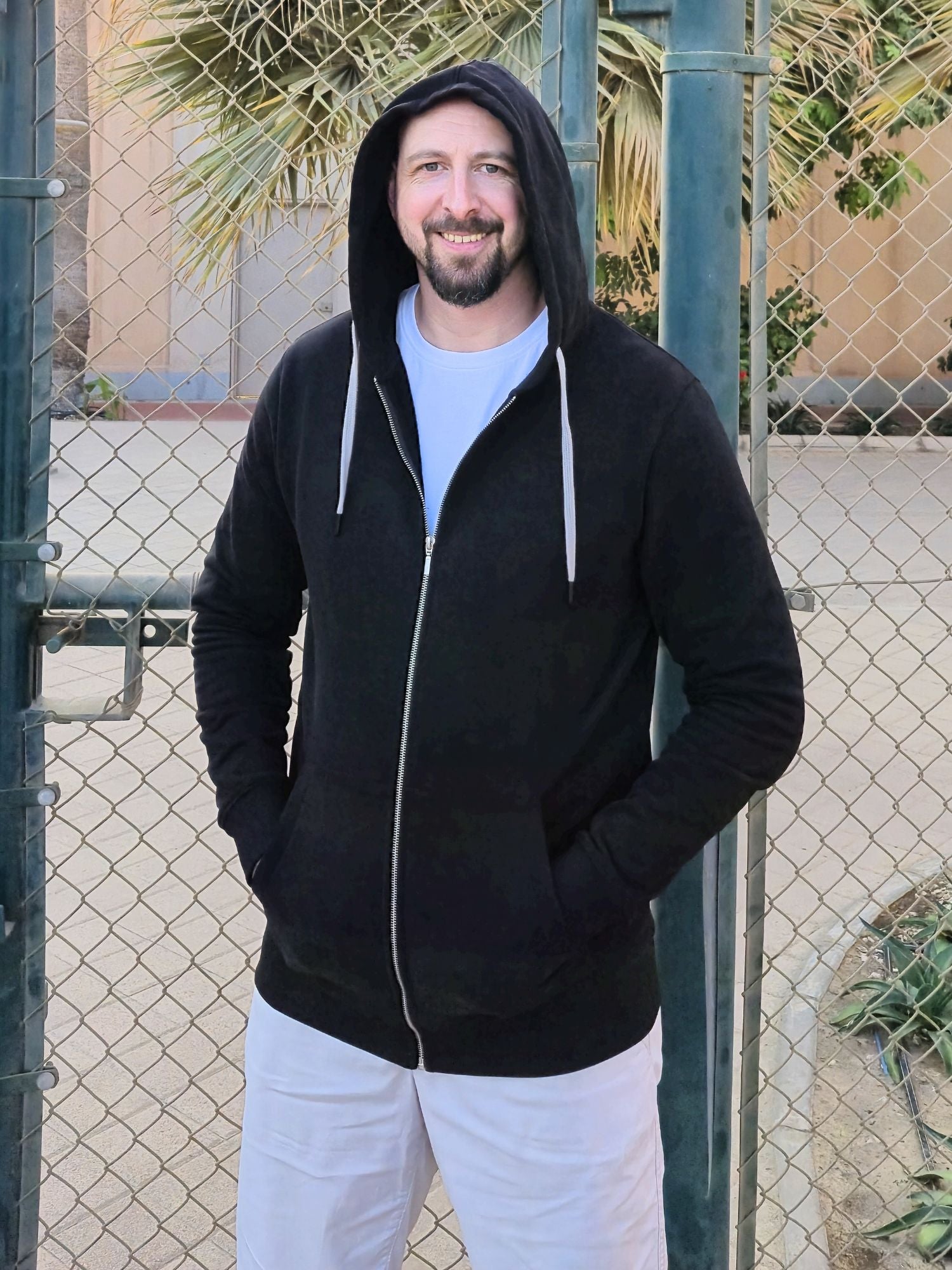 A tall slim guy wearing an XL tall black hoodie with hands in pockets and hood on, standing in a tennis court.