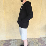 A side-on view of a tall and athletic guy wearing an XL tall black hoodie, standing in front of a beige wall, making hands in pockets and smiling.