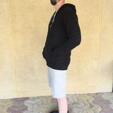 A side-on view of a tall and athletic guy wearing an XL tall black hoodie, standing in front of a beige wall, making hands in pockets and smiling.